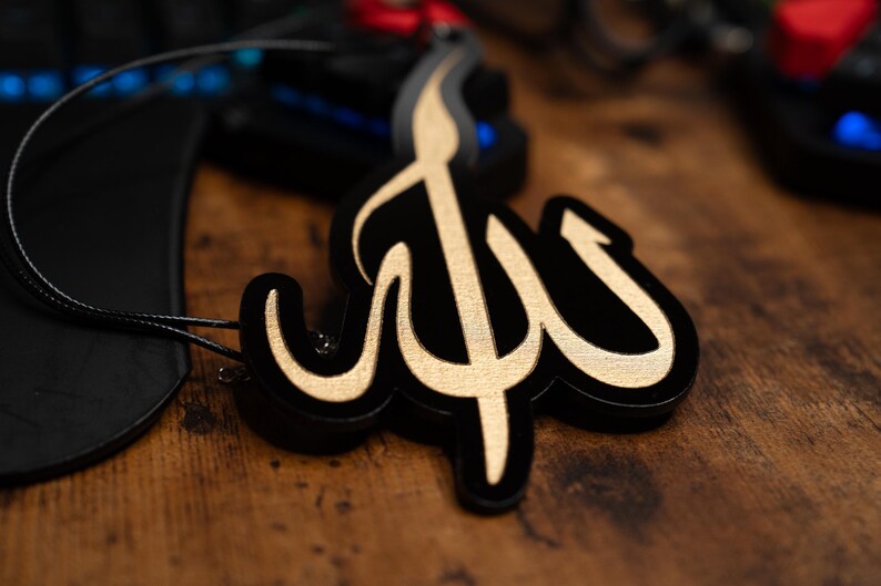 May include: A black and gold decorative ornament shaped like the Arabic word for God, "Allah". The ornament is hanging from a black cord.