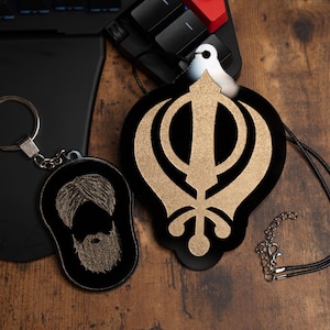 May include: Two black keychains with gold accents. One keychain features a Sikh symbol, the Khanda, and the other keychain features a bearded man wearing a turban.