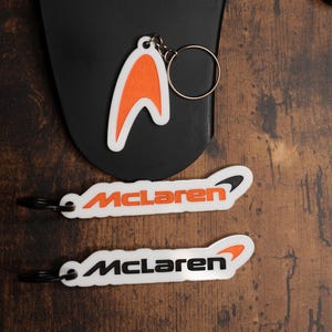 May include: Two white keychains with the word "McLaren" in black lettering and an orange McLaren logo. The keychains are on a brown wooden surface.