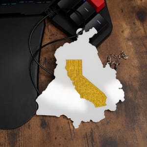 May include: A white outline of the state of California with a gold interior, hanging from a black cord and silver chain. The state outline is hanging on a wooden surface next to a black keyboard.