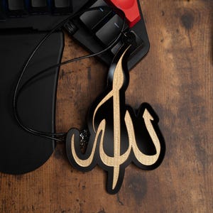 May include: A black and gold car air freshener with the Arabic word "Allah" written in a decorative script.