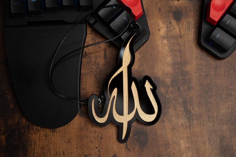 May include: A black and gold car air freshener with the Arabic word "Allah" written in a decorative script.