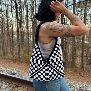May include: A black and white crochet bag with a geometric pattern. The bag has a black strap and a white lining. The bag is made of a soft, textured yarn and is a stylish accessory.