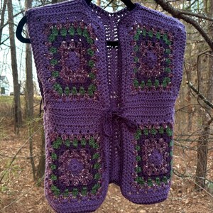 May include: A handmade, sleeveless crochet vest in shades of purple, green, and burgundy. The vest features a granny square design with a tie closure at the waist. The vest is hanging on a black hanger against a blurred outdoor background.