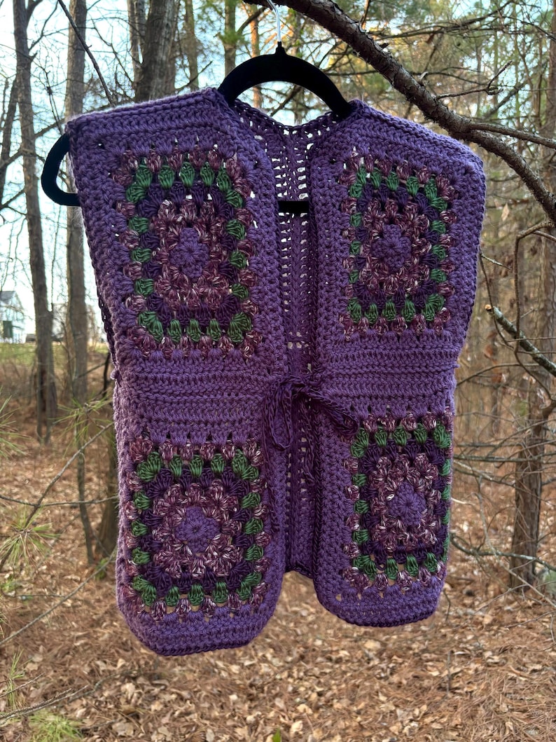 May include: A handmade, sleeveless crochet vest in shades of purple, green, and burgundy. The vest features a granny square design with a tie closure at the waist. The vest is hanging on a black hanger against a blurred outdoor background.