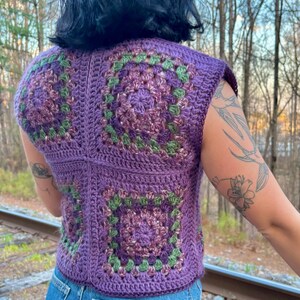 May include: A sleeveless, purple crochet top with square granny stitch patterns in shades of purple and green. The top is paired with blue denim jeans. The image is taken outdoors with a blurred background of trees and railroad tracks.