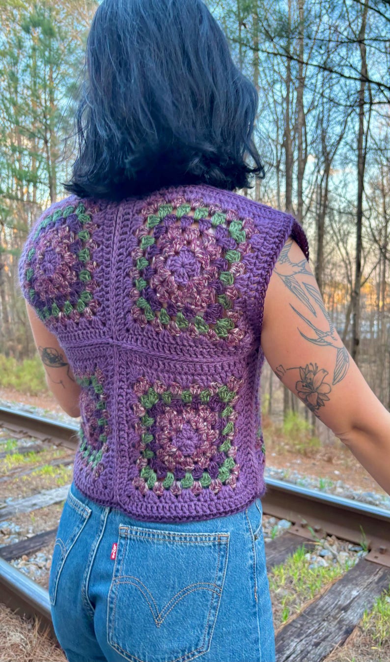 May include: A sleeveless, purple crochet top with square granny stitch patterns in shades of purple and green. The top is paired with blue denim jeans. The image is taken outdoors with a blurred background of trees and railroad tracks.