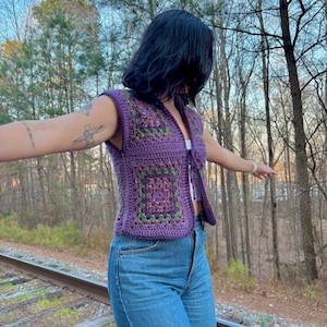 May include: A purple crochet vest with square floral patterns in shades of green and purple. The vest is sleeveless and has a tie closure. The person is wearing blue jeans.