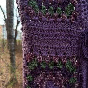 May include: Close-up of a handmade crochet item in shades of purple, green, and white. The textured pattern features a repeating floral design. The item appears to be a shawl or wrap, with a detailed, openwork stitch.