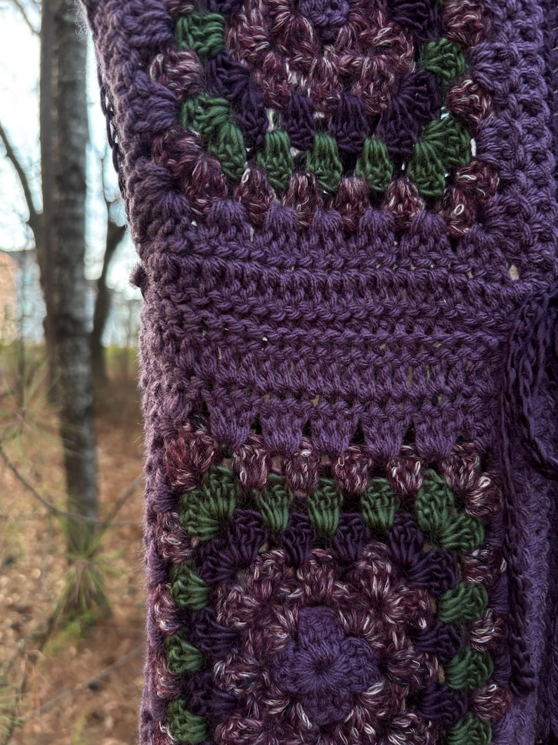 May include: Close-up of a handmade crochet item in shades of purple, green, and white. The textured pattern features a repeating floral design. The item appears to be a shawl or wrap, with a detailed, openwork stitch.