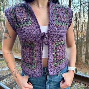 May include: A sleeveless, open-front crochet vest in shades of purple, green, and brown. The vest features a square pattern design and a tie closure at the chest. The garment is worn over a white top and paired with blue jeans.