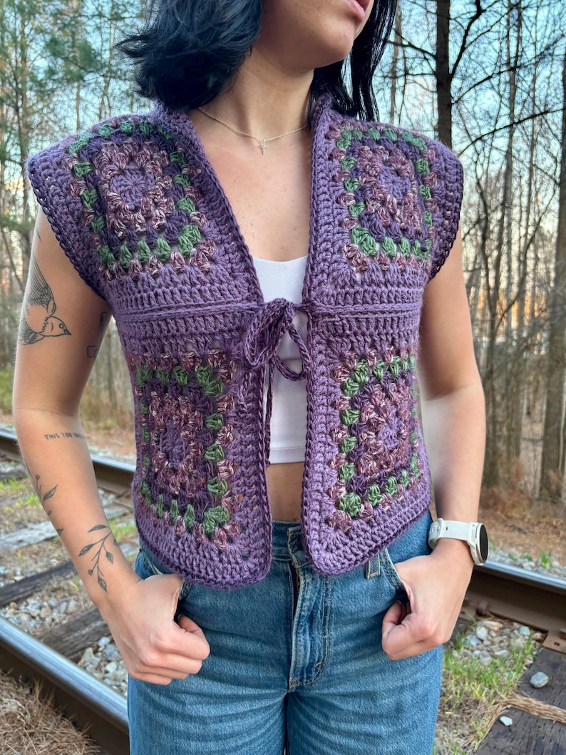 May include: A sleeveless, open-front crochet vest in shades of purple, green, and brown. The vest features a square pattern design and a tie closure at the chest. The garment is worn over a white top and paired with blue jeans.