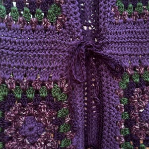 May include: Close-up of a handmade crochet cardigan in shades of purple, green, and white. The open-front cardigan features a tie closure and a granny square pattern. The texture of the yarn is visible.