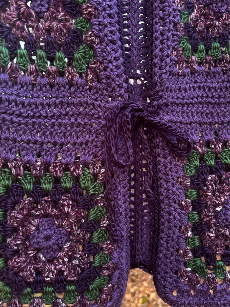 May include: Close-up of a handmade crochet cardigan in shades of purple, green, and white. The open-front cardigan features a tie closure and a granny square pattern. The texture of the yarn is visible.