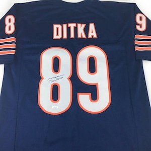 K&ouml;nnte beinhalten: Ein marineblaues Ditka-Football-Trikot mit wei&szlig;en Zahlen und Buchstaben. Die Nummer 89 ist auf der Vorder- und R&uuml;ckseite zu sehen, mit dem Namen "DITKA" &uuml;ber den Schultern. Orangefarbene Akzente und eine Unterschrift sind ebenfalls sichtbar.