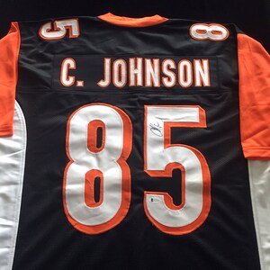 May include: Black and orange football jersey with the number 85 and the name "C. JOHNSON" on the back. The jersey is signed in black marker.