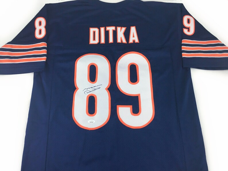 K&ouml;nnte beinhalten: Ein marineblaues Ditka-Football-Trikot mit wei&szlig;en Zahlen und Buchstaben. Die Nummer 89 ist auf der Vorder- und R&uuml;ckseite zu sehen, mit dem Namen "DITKA" &uuml;ber den Schultern. Orangefarbene Akzente und eine Unterschrift sind ebenfalls sichtbar.