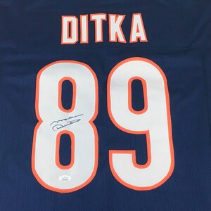 K&ouml;nnte beinhalten: Ein marineblaues Football-Trikot mit dem Namen "DITKA" in wei&szlig;en Buchstaben mit orangefarbenem Rand &uuml;ber den Schultern. Die Nummer "89" ist prominent in Wei&szlig; mit orangefarbenem Rand dargestellt. Das Trikot hat eine Unterschrift.