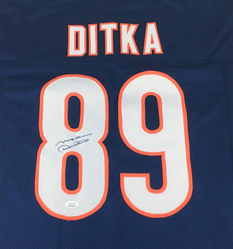 K&ouml;nnte beinhalten: Ein marineblaues Football-Trikot mit dem Namen "DITKA" in wei&szlig;en Buchstaben mit orangefarbenem Rand &uuml;ber den Schultern. Die Nummer "89" ist prominent in Wei&szlig; mit orangefarbenem Rand dargestellt. Das Trikot hat eine Unterschrift.