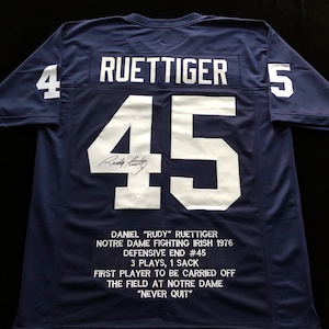 May include: A navy blue football jersey with the number 45 on the back. The name "RUETTIGER" is above the number and the number 4 is on the left sleeve and the number 5 is on the right sleeve. The jersey is signed by "Rudy Ruettiger". The text "DANIEL "RUDY" RUETTIGER NOTRE DAME FIGHTING IRISH 1976 DEFENSIVE END #45 3 PLAYS, 1 SACK FIRST PLAYER TO BE CARRIED OFF THE FIELD AT NOTRE DAME "NEVER QUIT" is printed on the back of the jersey.
