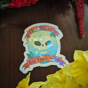 May include: A holographic sticker with a skull and crossbones design. The sticker says "The Bones Are Their Money".