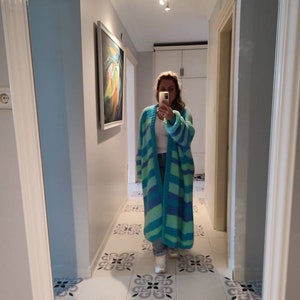May include: A long, open-front cardigan in shades of teal and blue, with horizontal stripes. The cardigan is knee-length and appears to be made of a chunky knit material. The person is wearing jeans and white sneakers.