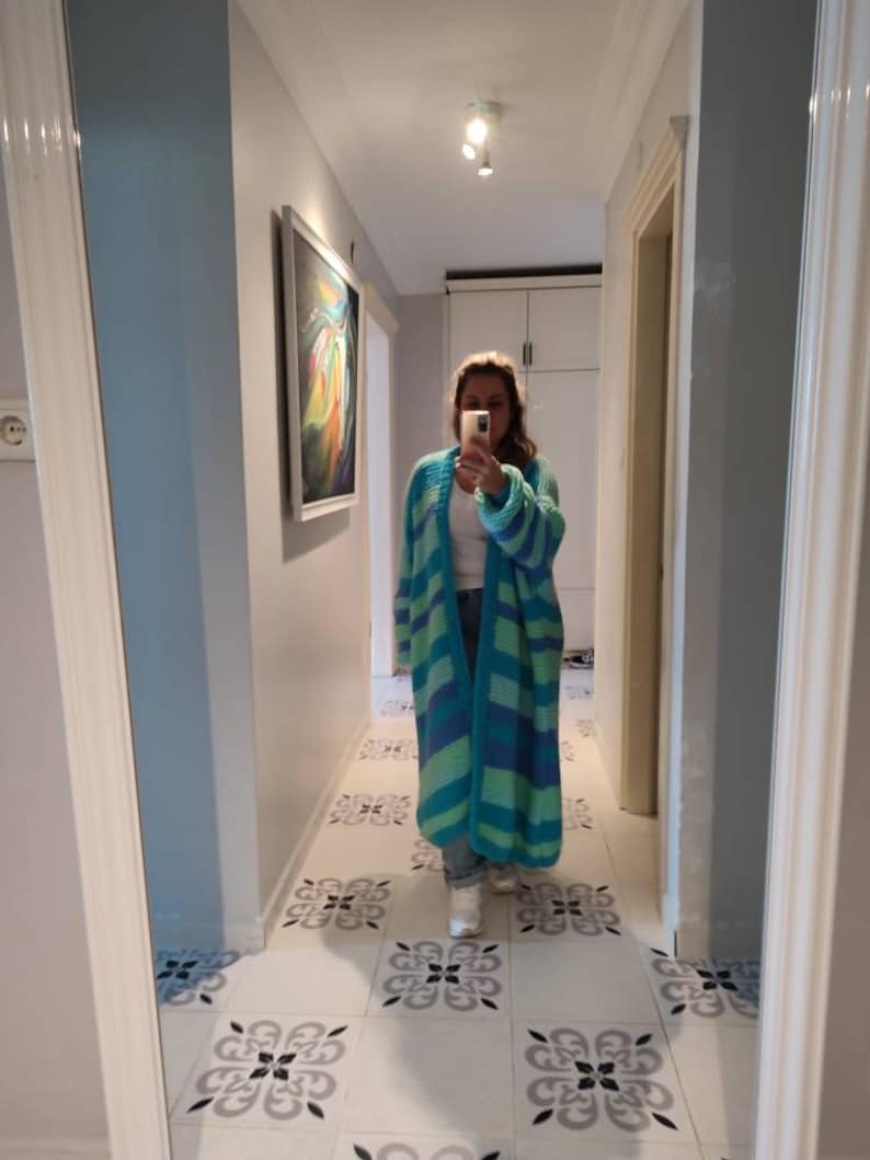 May include: A long, open-front cardigan in shades of teal and blue, with horizontal stripes. The cardigan is knee-length and appears to be made of a chunky knit material. The person is wearing jeans and white sneakers.