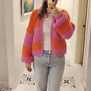 May include: A chunky knit cardigan featuring wide horizontal stripes in vibrant orange and bright pink. The garment has long sleeves, a ribbed hem, and a ribbed front opening. It is worn over a plain white top and light blue denim jeans.