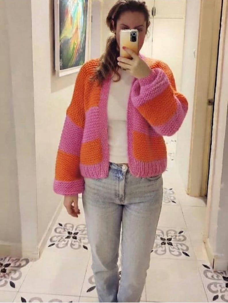 May include: A chunky knit cardigan featuring wide horizontal stripes in vibrant orange and bright pink. The garment has long sleeves, a ribbed hem, and a ribbed front opening. It is worn over a plain white top and light blue denim jeans.