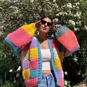 May include: A colorful, patchwork knit cardigan in shades of pink, yellow, blue, and purple. The chunky knit sweater has a relaxed fit with long sleeves. The cardigan is open-front and appears to be handmade, perfect for a cozy, stylish look.