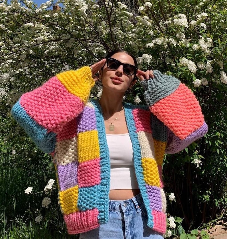 May include: A colorful, patchwork knit cardigan in shades of pink, yellow, blue, and purple. The chunky knit sweater has a relaxed fit with long sleeves. The cardigan is open-front and appears to be handmade, perfect for a cozy, stylish look.