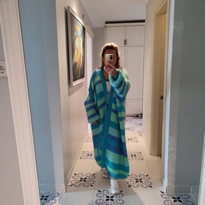 May include: A long, oversized, striped cardigan in shades of blue and green. The cardigan is made of a chunky knit material and has a relaxed fit. The person is wearing white sneakers.