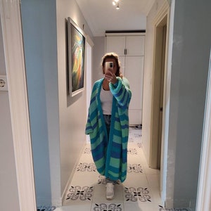 May include: A long, open-front cardigan in shades of turquoise and blue, with wide horizontal stripes. The chunky knitwear is worn over a white top and jeans. The person is standing in a hallway with a decorative tile floor.