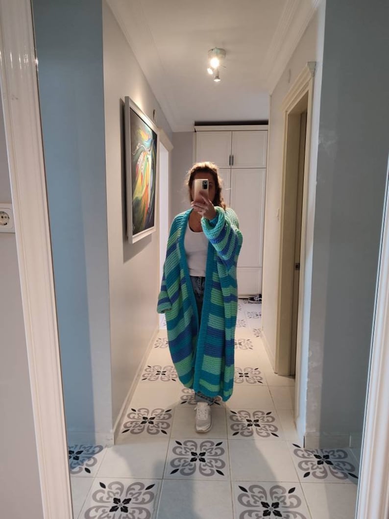 May include: A long, open-front cardigan in shades of turquoise and blue, with wide horizontal stripes. The chunky knitwear is worn over a white top and jeans. The person is standing in a hallway with a decorative tile floor.