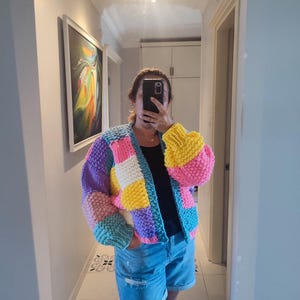 May include: A colorful, chunky knit cardigan in a patchwork design. The sweater features squares of pink, yellow, purple, blue, and white. The sleeves are a puffy style. The person in the image is wearing denim shorts and a black top.