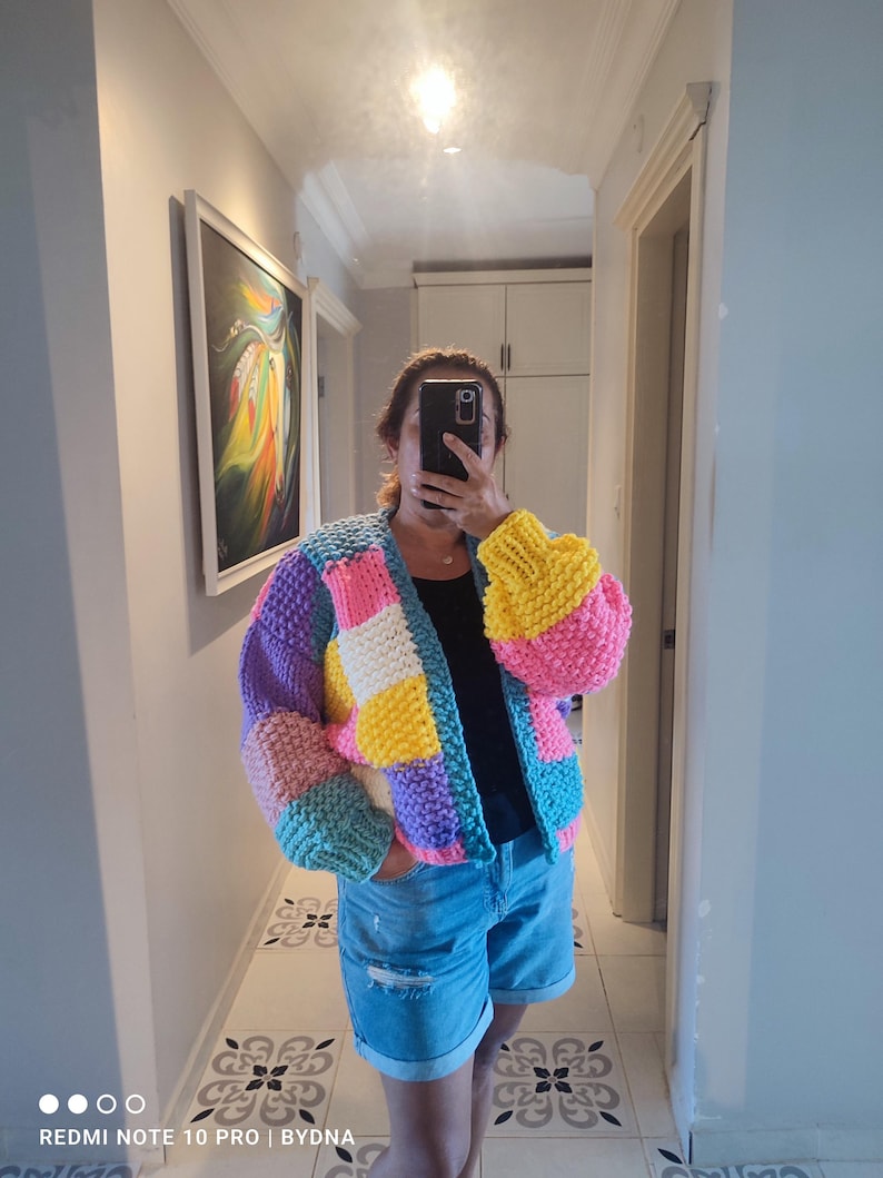 May include: A colorful, chunky knit cardigan in a patchwork design. The sweater features squares of pink, yellow, purple, blue, and white. The sleeves are a puffy style. The person in the image is wearing denim shorts and a black top.