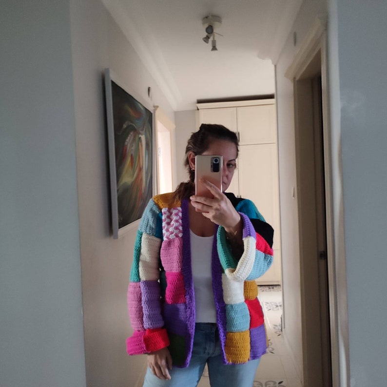 Puede incluir: Un c&aacute;rdigan de crochet colorido con un dise&ntilde;o de patchwork en tonos de rosa, morado, azul, verde, amarillo y naranja. El c&aacute;rdigan tiene una abertura larga en la parte delantera y mangas largas.