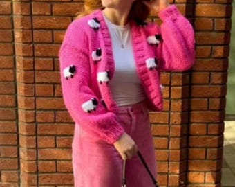 Könnte beinhalten: Ein leuchtend pinkfarbener Cardigan mit weißen und schwarzen Schafapplikationen. Der offen geschnittene Pullover hat lange Ärmel und wird mit einer pinkfarbenen Hose getragen. Das Outfit wird von einer Person vor einer Backsteinmauer getragen.