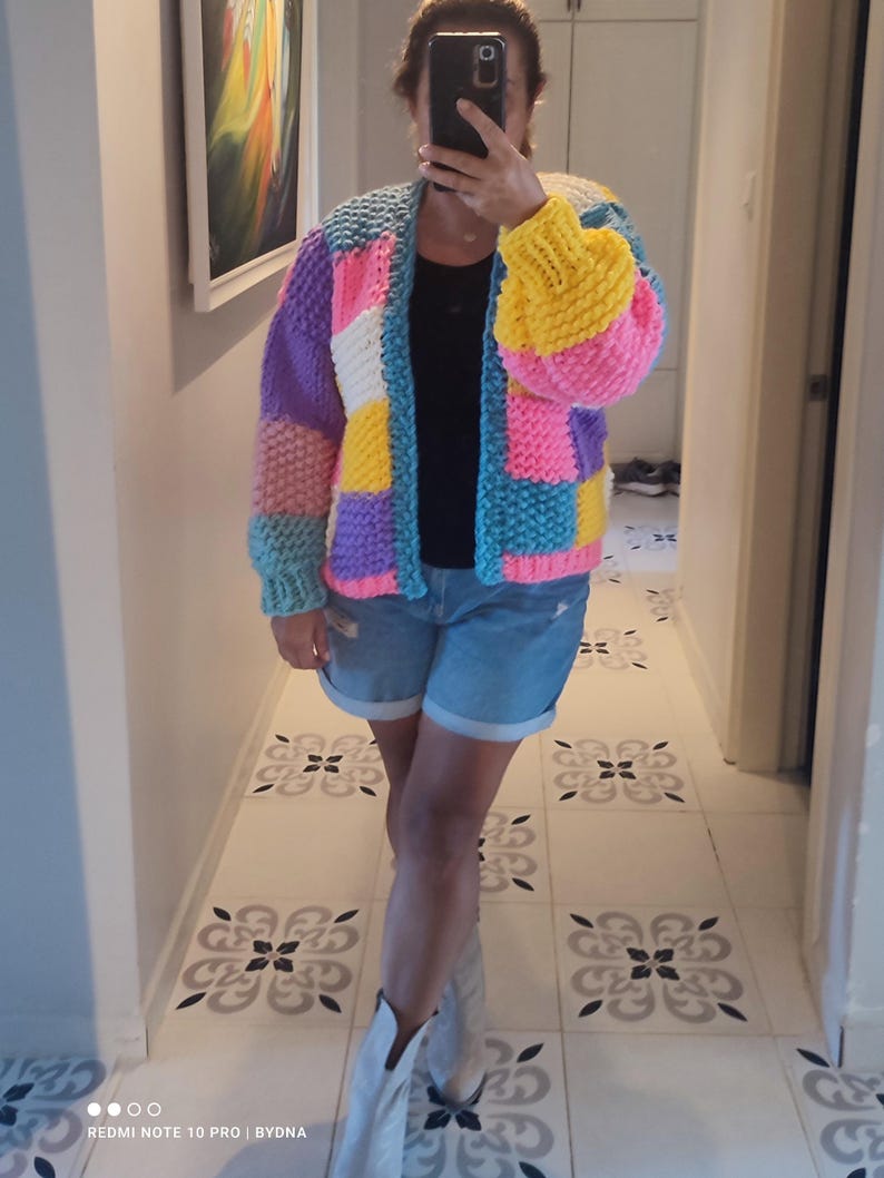 May include: A colorful, patchwork knit cardigan in shades of pink, yellow, blue, purple, and white. The open-front sweater has long sleeves and a chunky knit texture. The person in the image is wearing denim shorts and white boots.