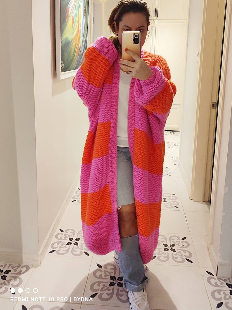 May include: A long, chunky knit cardigan featuring wide horizontal stripes in vibrant pink and bright orange. The garment has an open front, long sleeves with dropped shoulders, and a thick ribbed trim along the collar and front edges.