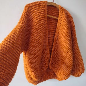 Puede incluir: Un c&aacute;rdigan de punto grueso en un tono naranja vibrante, con un dise&ntilde;o frontal abierto, mangas largas y holgadas, y un patr&oacute;n de punto texturizado. La prenda se muestra en una percha de madera, destacando su ajuste relajado y tejido grueso.