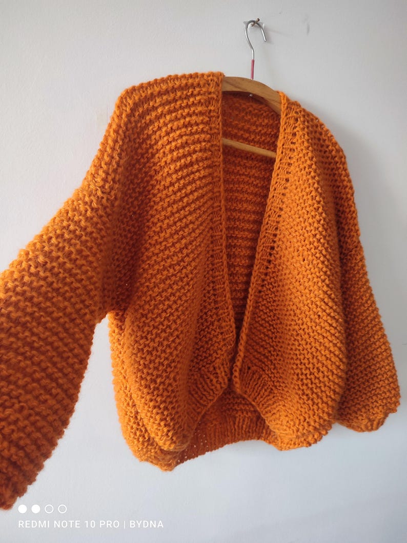 Puede incluir: Un c&aacute;rdigan de punto grueso en un tono naranja vibrante, con un dise&ntilde;o frontal abierto, mangas largas y holgadas, y un patr&oacute;n de punto texturizado. La prenda se muestra en una percha de madera, destacando su ajuste relajado y tejido grueso.