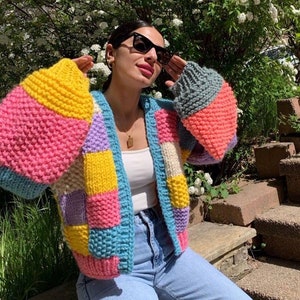 May include: A colorful, patchwork knit cardigan in shades of pink, yellow, blue, and gray. The open-front sweater has wide sleeves and is worn with a white top and light blue jeans. The cardigan is a cozy and stylish outerwear option.