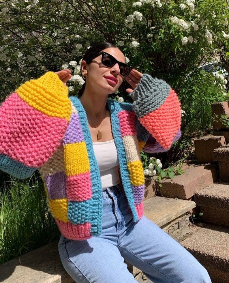 May include: A colorful, patchwork knit cardigan in shades of pink, yellow, blue, and gray. The open-front sweater has wide sleeves and is worn with a white top and light blue jeans. The cardigan is a cozy and stylish outerwear option.
