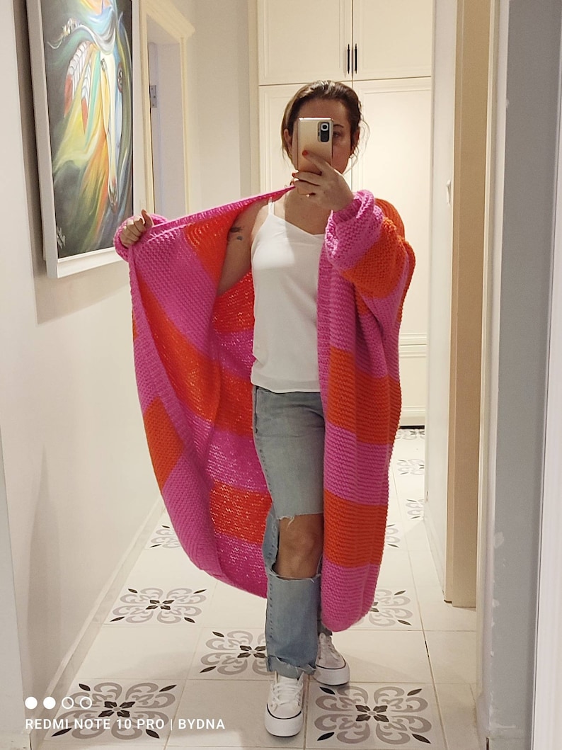 May include: A long, chunky knit cardigan featuring wide horizontal stripes in vibrant pink and bright orange. The open-front garment has long sleeves and a relaxed, oversized fit, showcasing a thick, textured yarn construction throughout the piece.