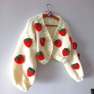 Peut inclure: Cardigan en tricot crème orné de fraises rouges avec des feuilles vertes. Le pull court a des manches longues et bouffantes et est suspendu à un cintre en bois. Une petite décoration florale est en haut du cintre.