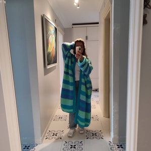 May include: A long, striped cardigan in shades of blue and green. The open-front sweater features wide horizontal stripes and is made of a chunky knit material. The cardigan is worn over a white top and jeans.