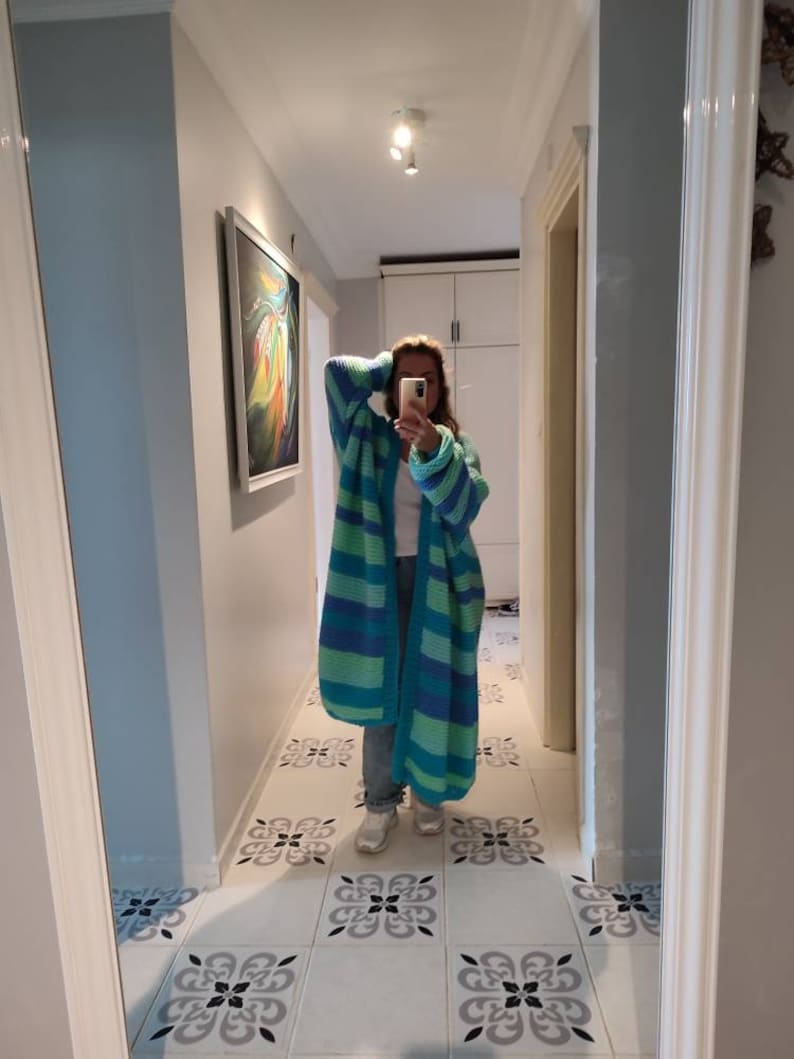 May include: A long, striped cardigan in shades of blue and green. The open-front sweater features wide horizontal stripes and is made of a chunky knit material. The cardigan is worn over a white top and jeans.