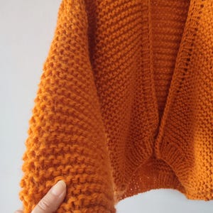 Puede incluir: Un c&aacute;rdigan de color naranja vibrante abierto por delante, con una textura de punto grueso, mangas largas y un dobladillo acanalado. La prenda est&aacute; confeccionada con hilo grueso, mostrando un patr&oacute;n de puntada consistente en todo el cuerpo.