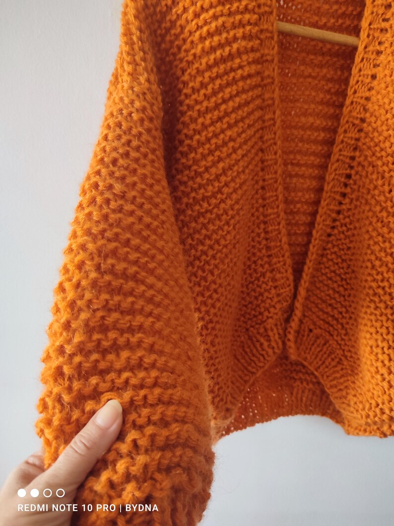 Puede incluir: Un c&aacute;rdigan de color naranja vibrante abierto por delante, con una textura de punto grueso, mangas largas y un dobladillo acanalado. La prenda est&aacute; confeccionada con hilo grueso, mostrando un patr&oacute;n de puntada consistente en todo el cuerpo.